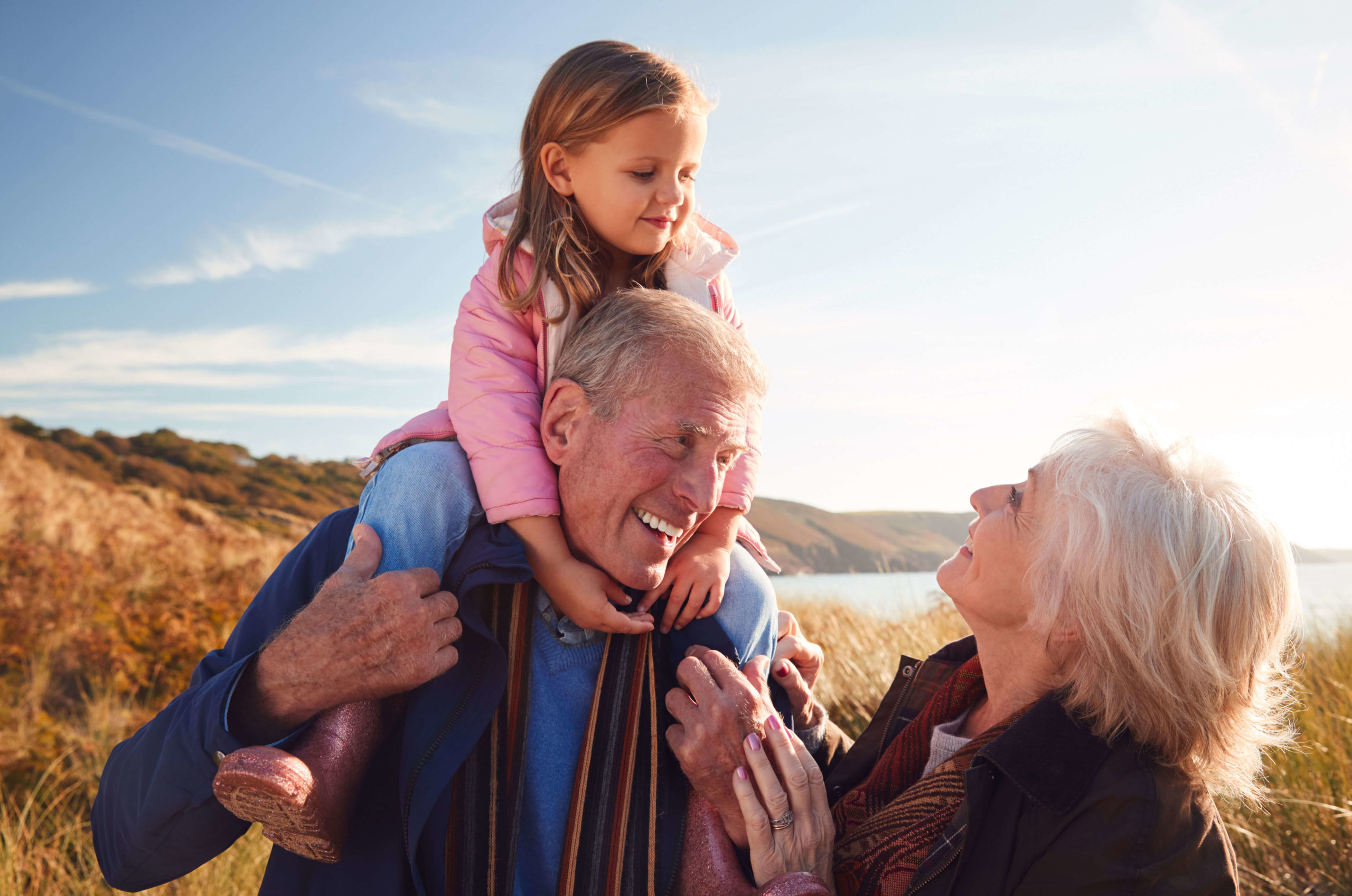Joyful multigenerational family moment with a grandfather carrying his granddaughter on his shoulders and the grandmother smiling at them, representing happy retirement and family values.